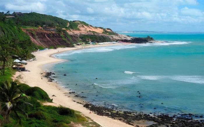 Vista panorámica de la playa en Pipa, Brasil, con acantilados rojizos, mar azul y vegetación tropical.