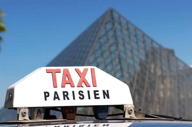 Taxi de París con cartel 'TAXI PARISIEN' en primer plano, frente a la pirámide del Louvre en París, bajo un cielo despejado.